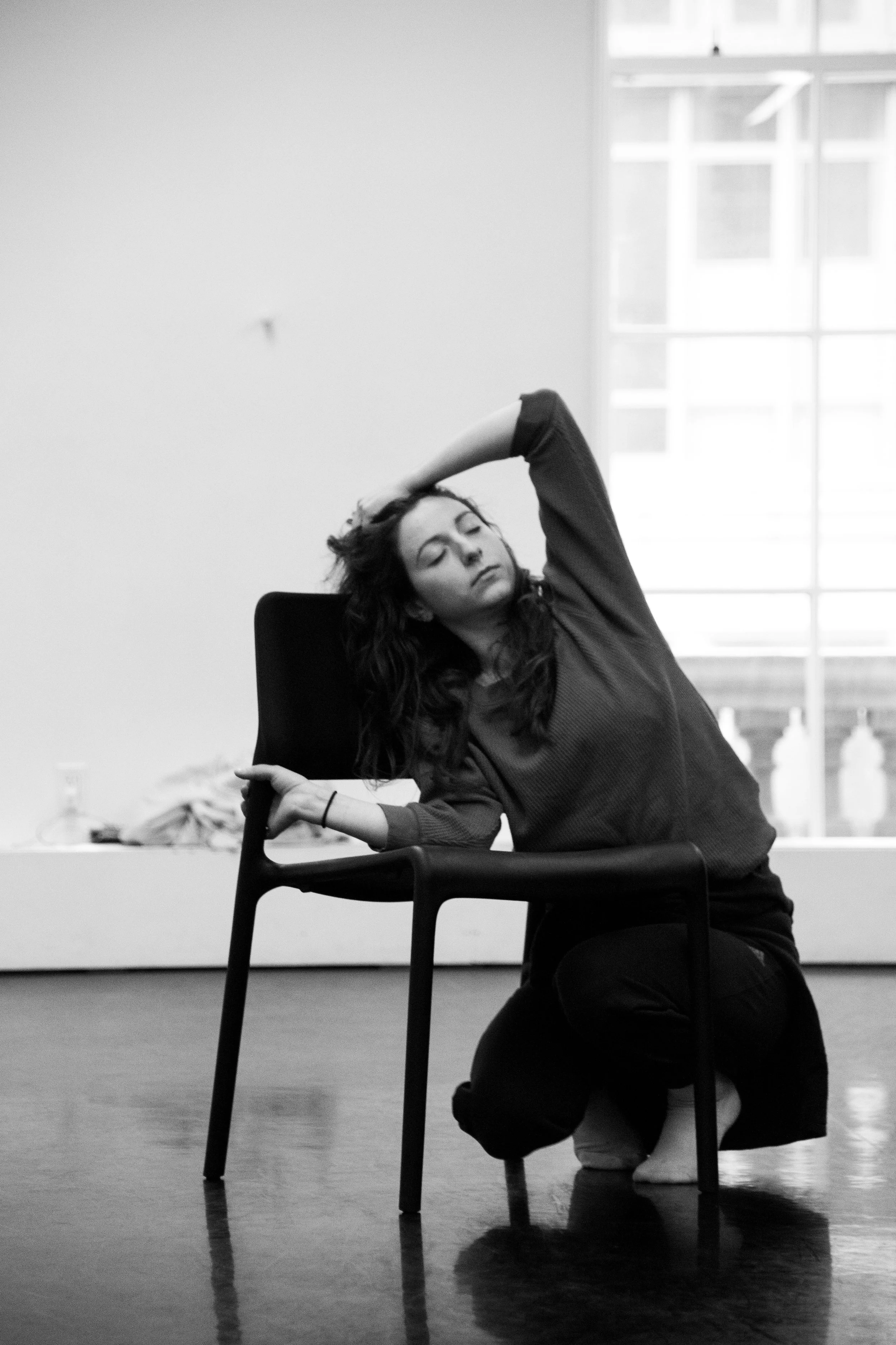 A woman with long curly hair sitting on the floor behind a black chair with a serious expression, her eyes closed, one arm raised over her head, in a room with large windows.