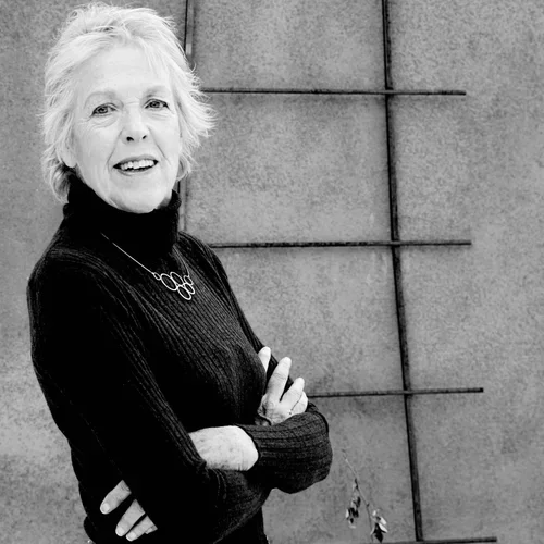 Black and white photo of an elderly woman with short, light-colored hair, smiling, wearing a dark turtleneck and a necklace, standing outdoors against a concrete wall.