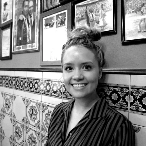 A young woman with a top knot hairstyle smiling at the camera, standing in front of a wall decorated with framed photographs and patterned tiles.