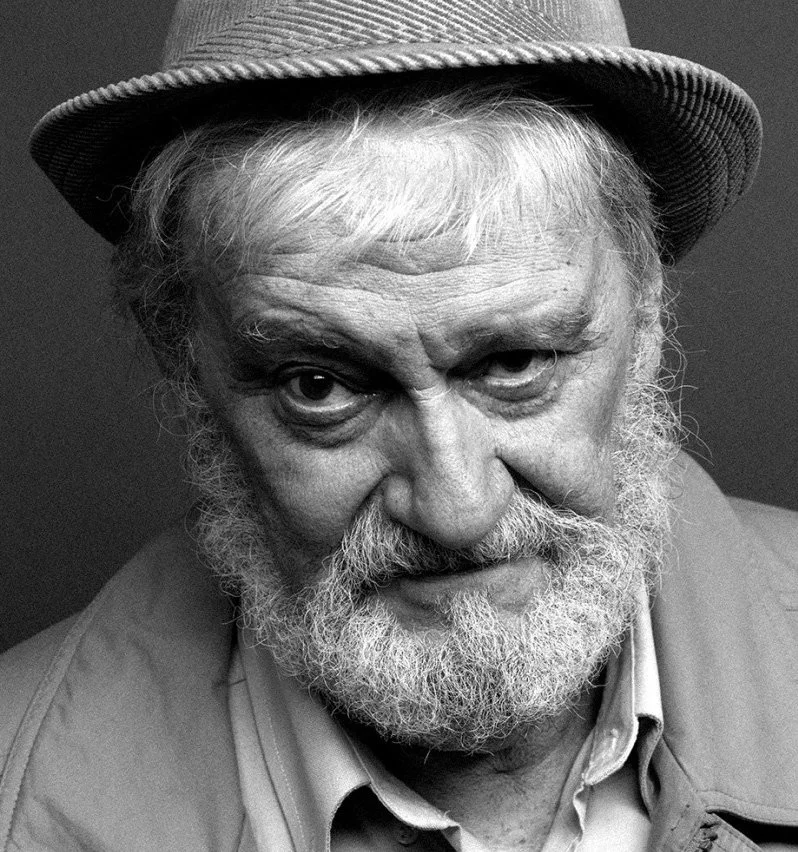 Black-and-white close-up portrait of an elderly man with a beard wearing a hat and collared shirt.