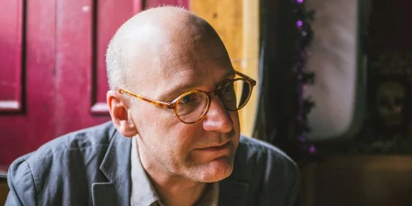 Close-up of a bald man with glasses, wearing a dark blazer, in an indoor setting with a red door and plants in the background.