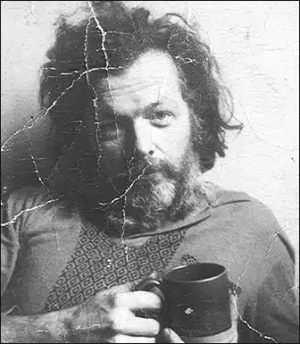 Black and white photo of a man with long, messy hair and a beard, holding a cup.