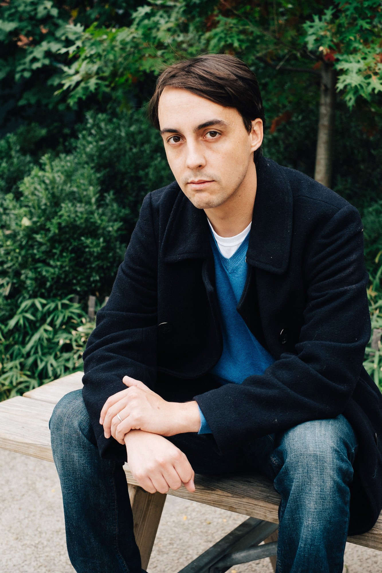 A man in a black coat and blue sweater sitting on a wooden bench outdoors surrounded by green bushes and trees.