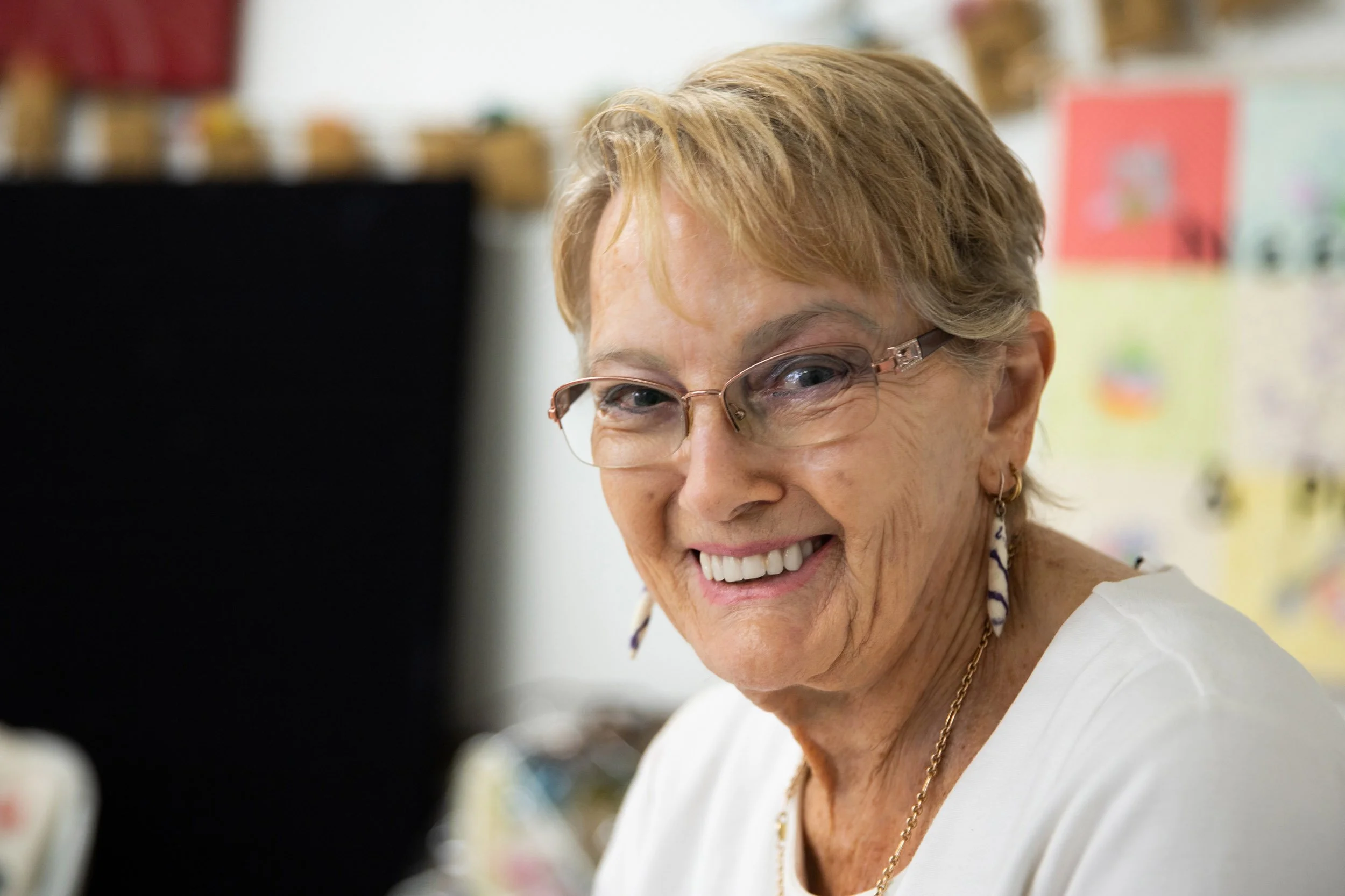 A smiling elderly woman with short, light brown hair and glasses, looking to the side, in an indoor setting with colorful decorations and drawings in the background.