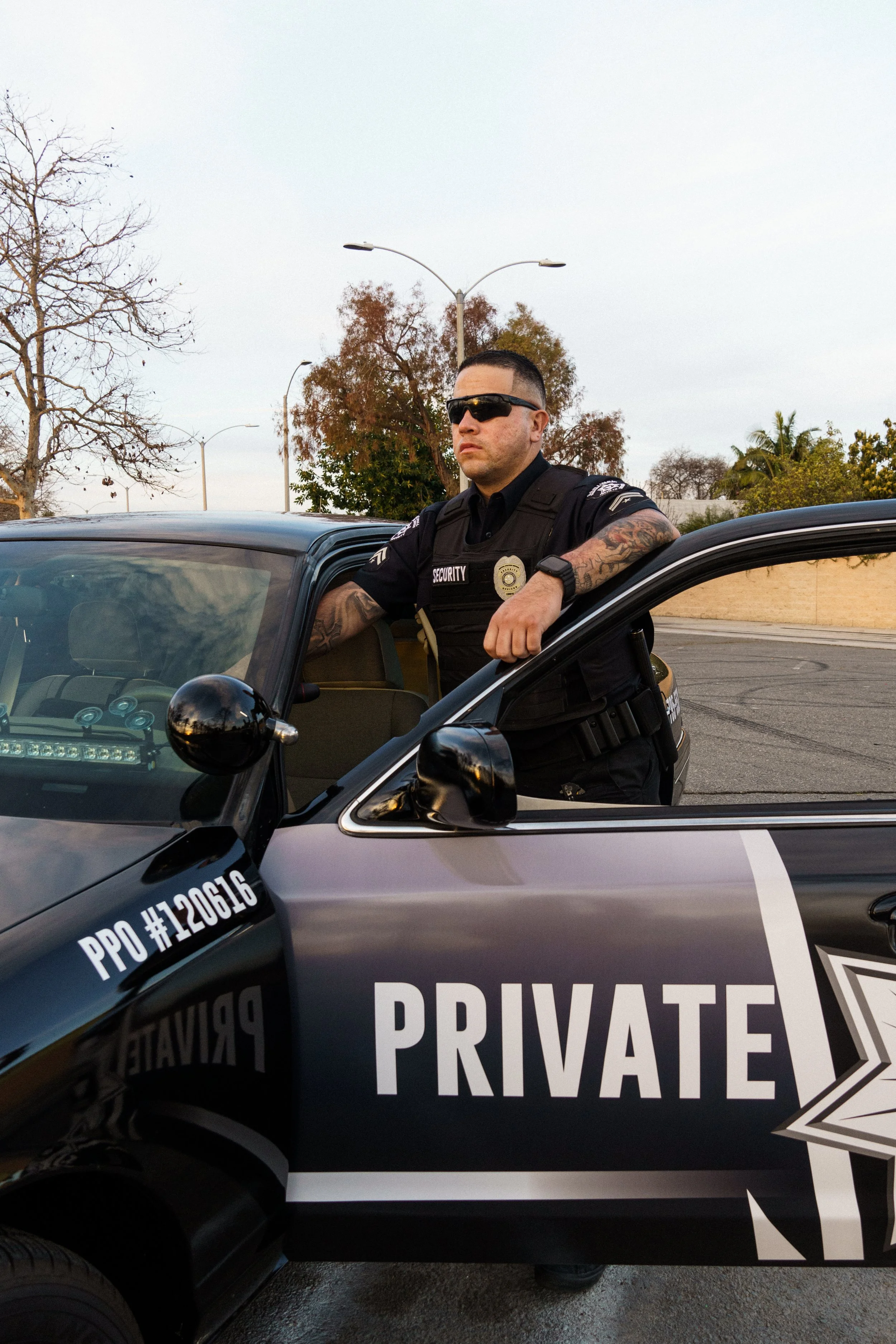 A security officer with tattoos, sunglasses, and a black uniform leaning on a black police car marked 'PRIVATE' and 'PPO #120616'.