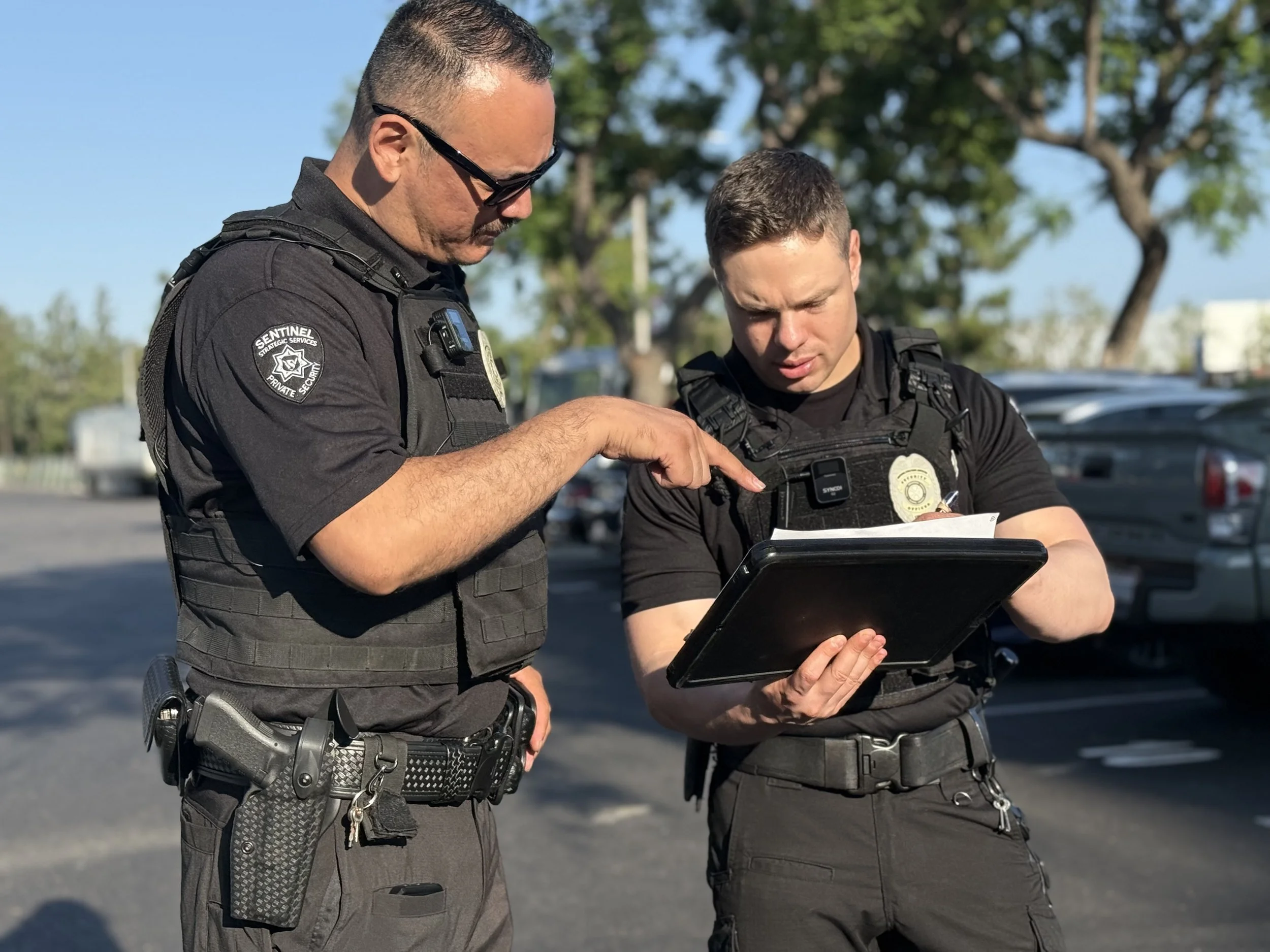 Two security officers in uniform are looking at a tablet device together in a parking lot, with trees and parked cars in the background.