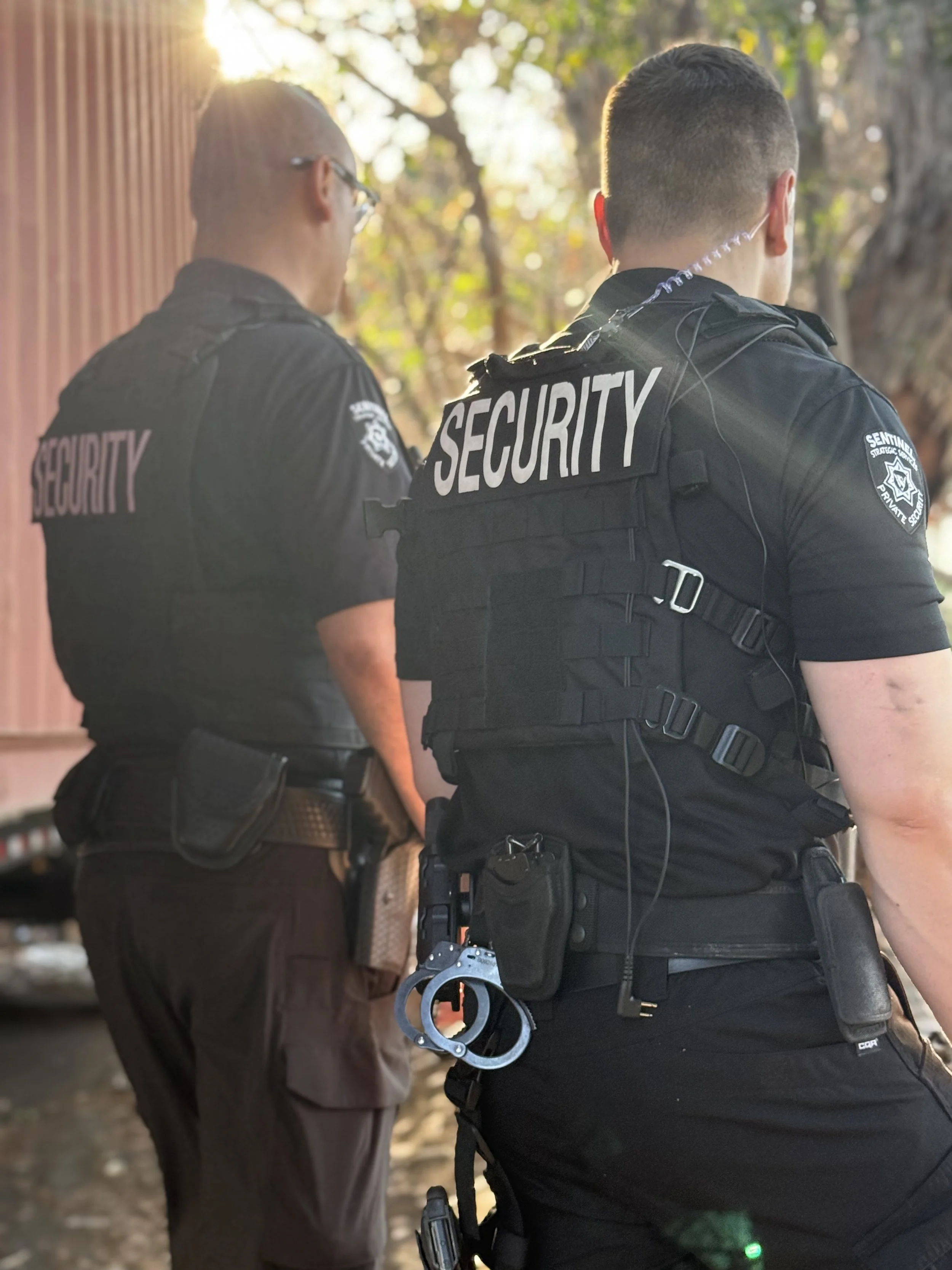 Two security officers standing outdoors, one with handcuffs attached to their belt.