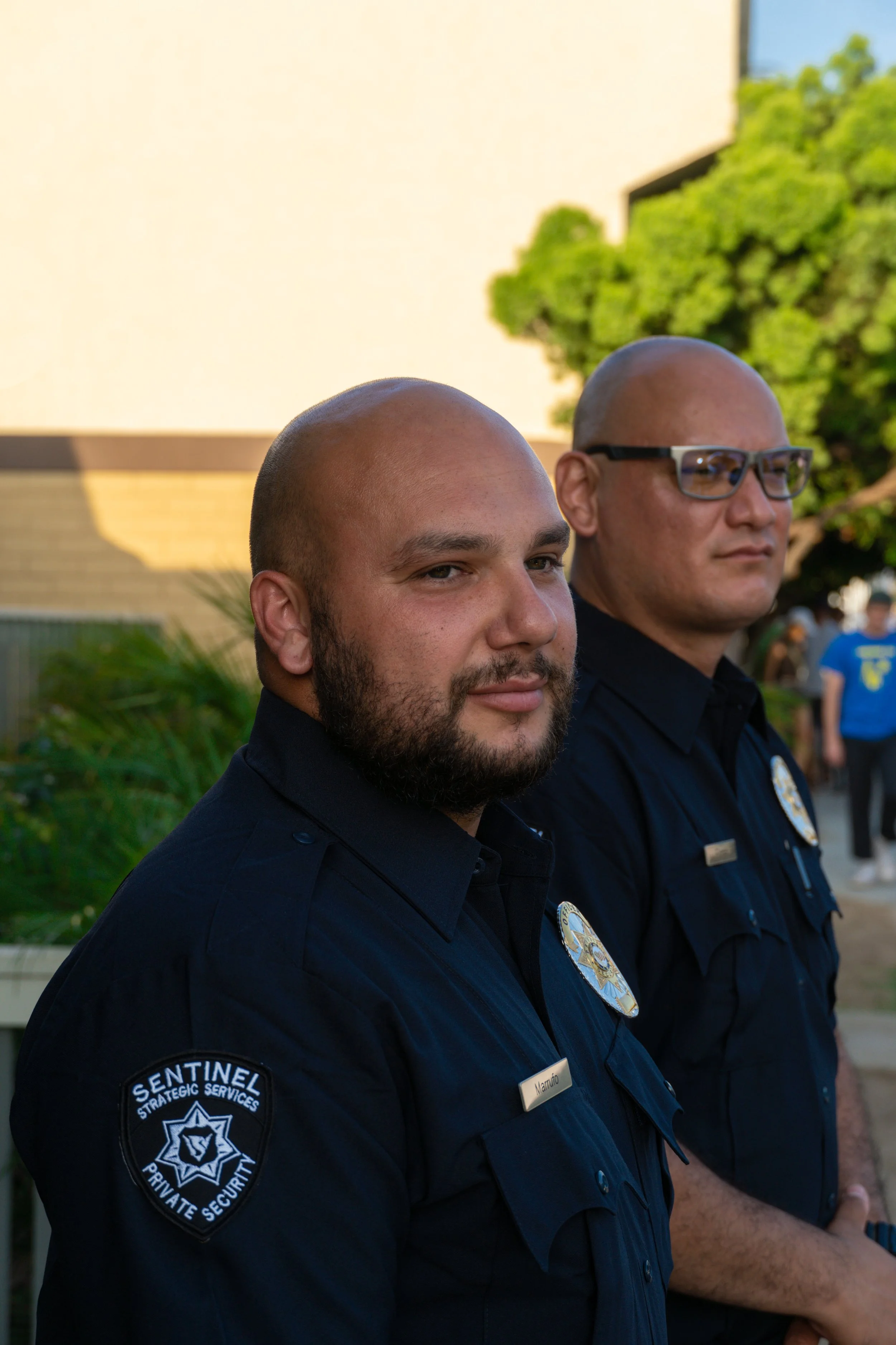 Two uniformed security officers, one with a badge and patch, standing outdoors with greenery and a building in the background.