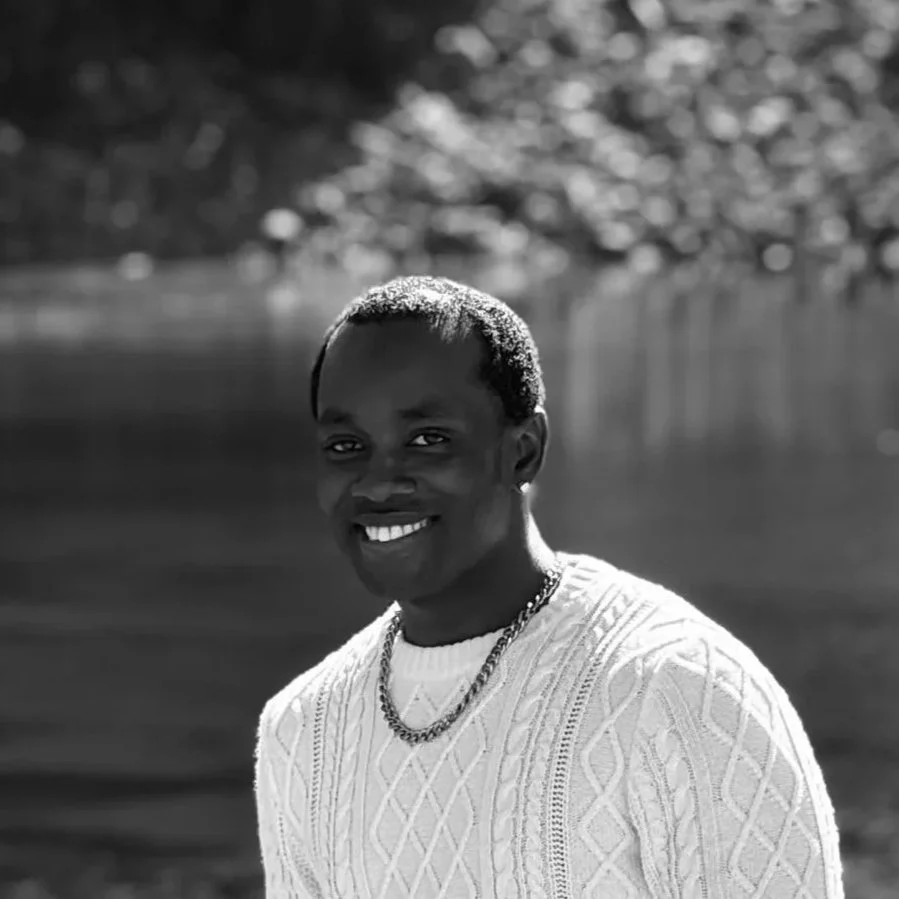 A young person with short hair, smiling, wearing a light color knitted sweater and a chain necklace, standing outdoors near a body of water.