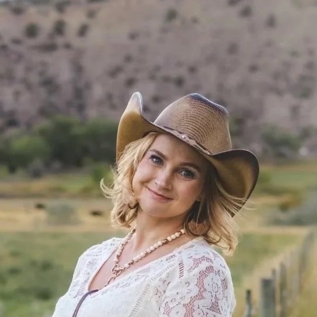 A woman in a light-colored lace top and a wide-brimmed cowboy hat, smiling outdoors with a blurred natural landscape in the background.