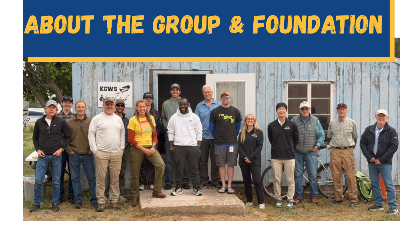 Group of 15 people standing in front of a weathered wooden building with a sign for KGWS. The group includes men and women dressed casually, some with jackets, and one with a bicycle. There are trees and a vehicle partially visible in the background.
