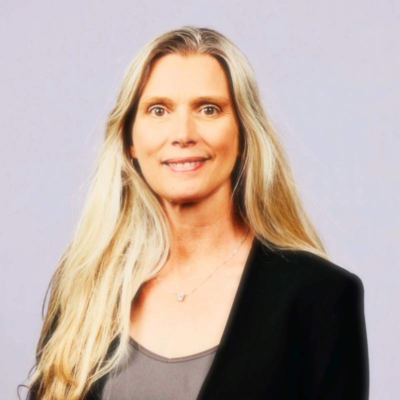 A woman with long blonde hair, smiling, wearing a black blazer over a gray top, against a plain light background.