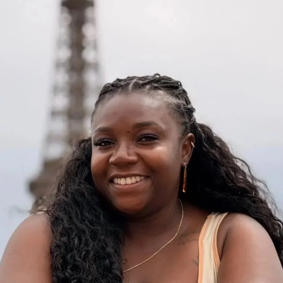 A smiling woman with dark curly hair wearing a sleeveless top, standing outdoors with a blurred tower or structure in the background.