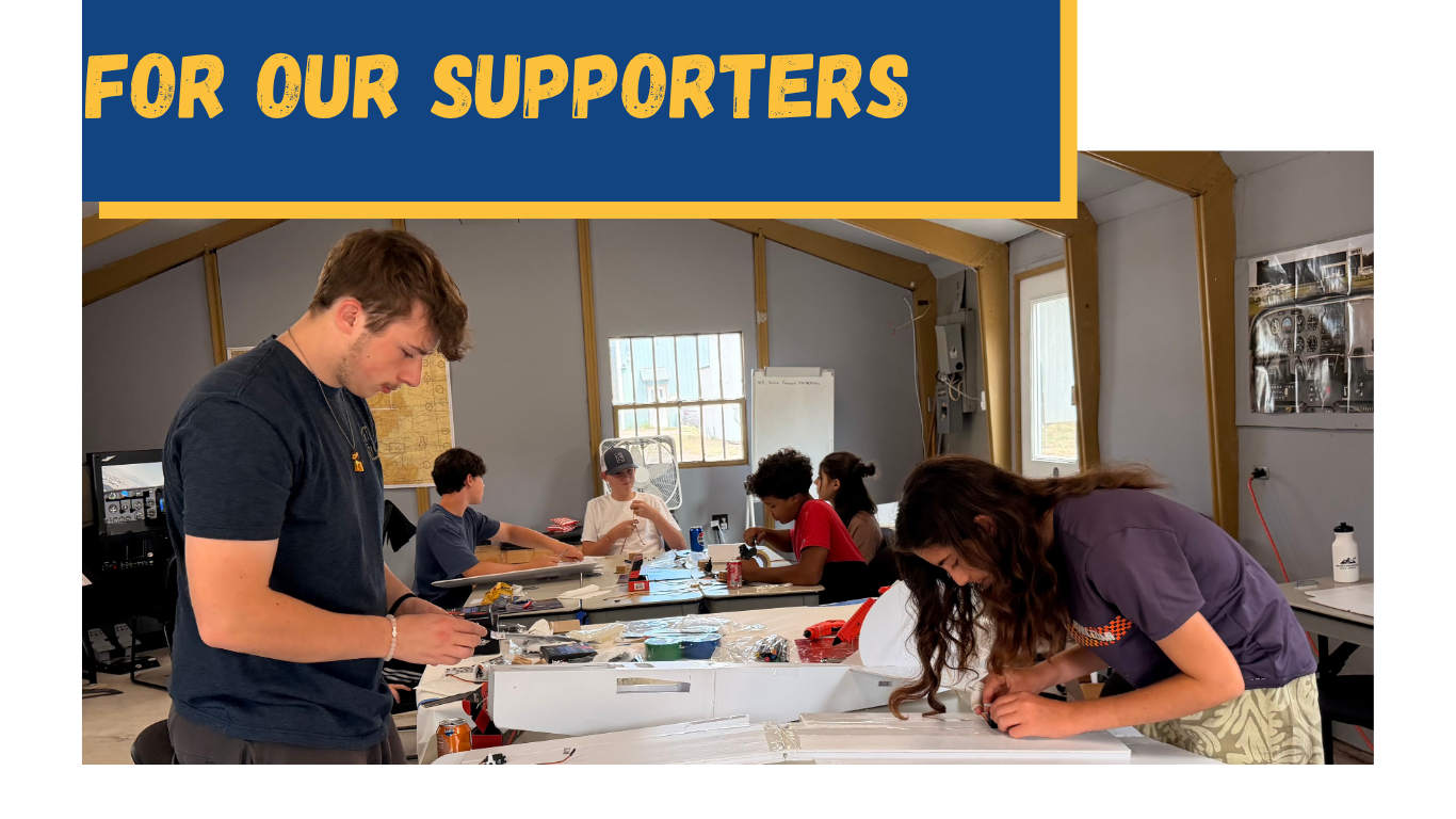 A group of young people working on projects at a workshop table inside a room with windows, with a banner at the top that reads 'For Our Supporters'.