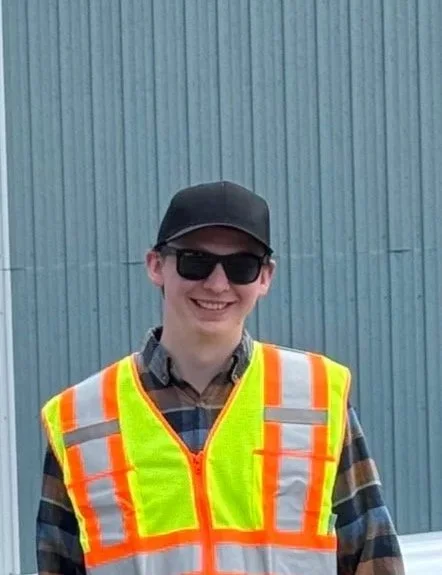 Young man wearing a safety vest, sunglasses, and a cap, smiling in front of a corrugated metal wall.