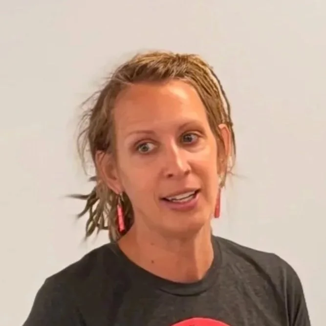 A woman with short, light brown hair wearing a black shirt and pink earrings, speaking in front of a plain light-colored wall.
