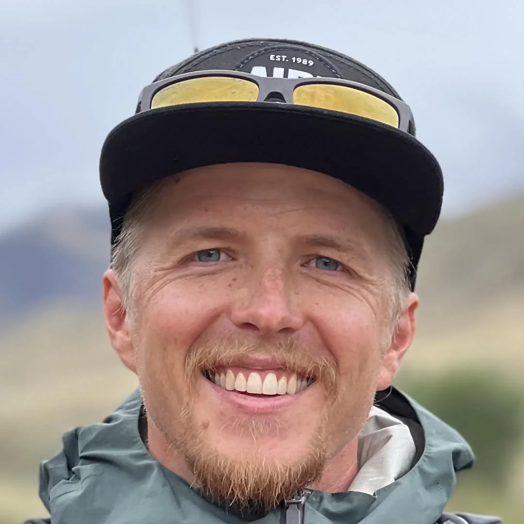 Close-up of a smiling man with blue eyes, wearing a black cap with sunglasses on top, and a gray jacket outdoors.