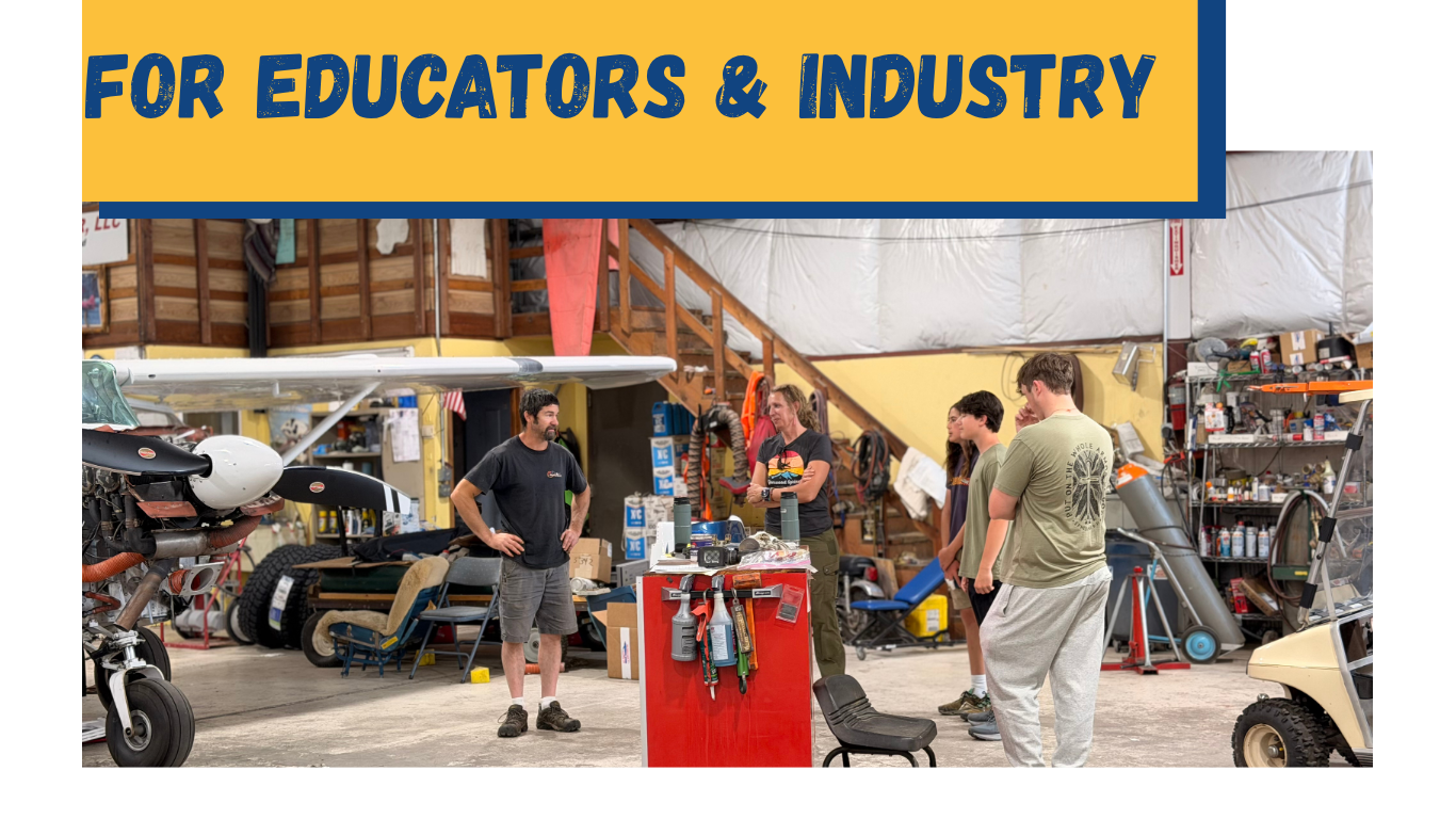 A group of five people inside a workshop or garage, standing around and talking. The workshop has various tools, equipment, and a small aircraft or drone visible.