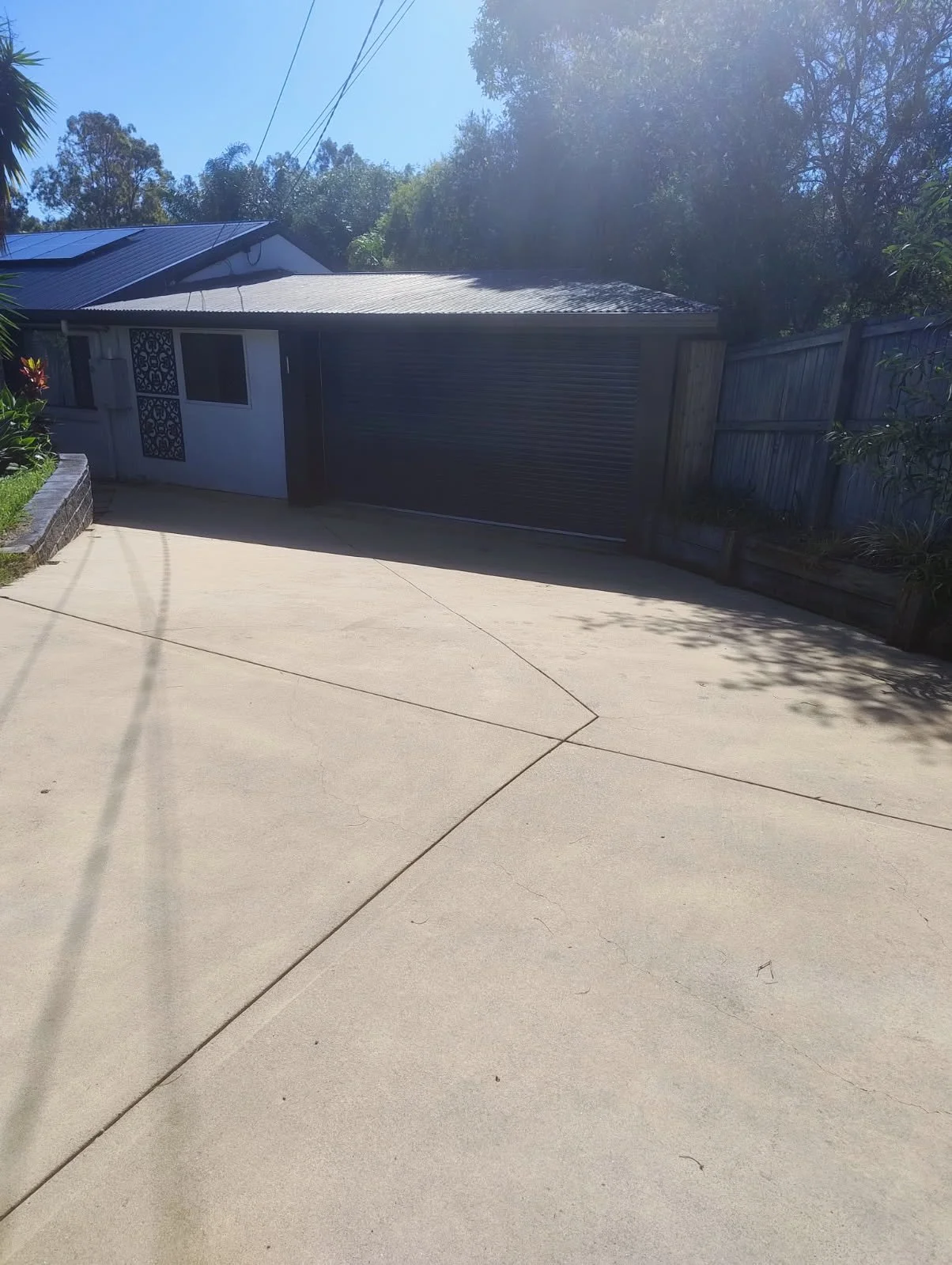 A driveway leading to a house with a closed garage door, surrounded by a wooden fence and some plants, with trees and a bright sky in the background.