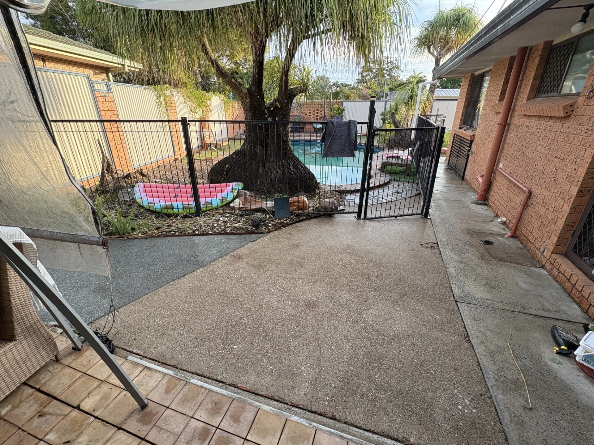 View of a backyard patio with a small enclosed kiddie pool and covered swimming pool, surrounded by a fence, with plants and trees in the background.