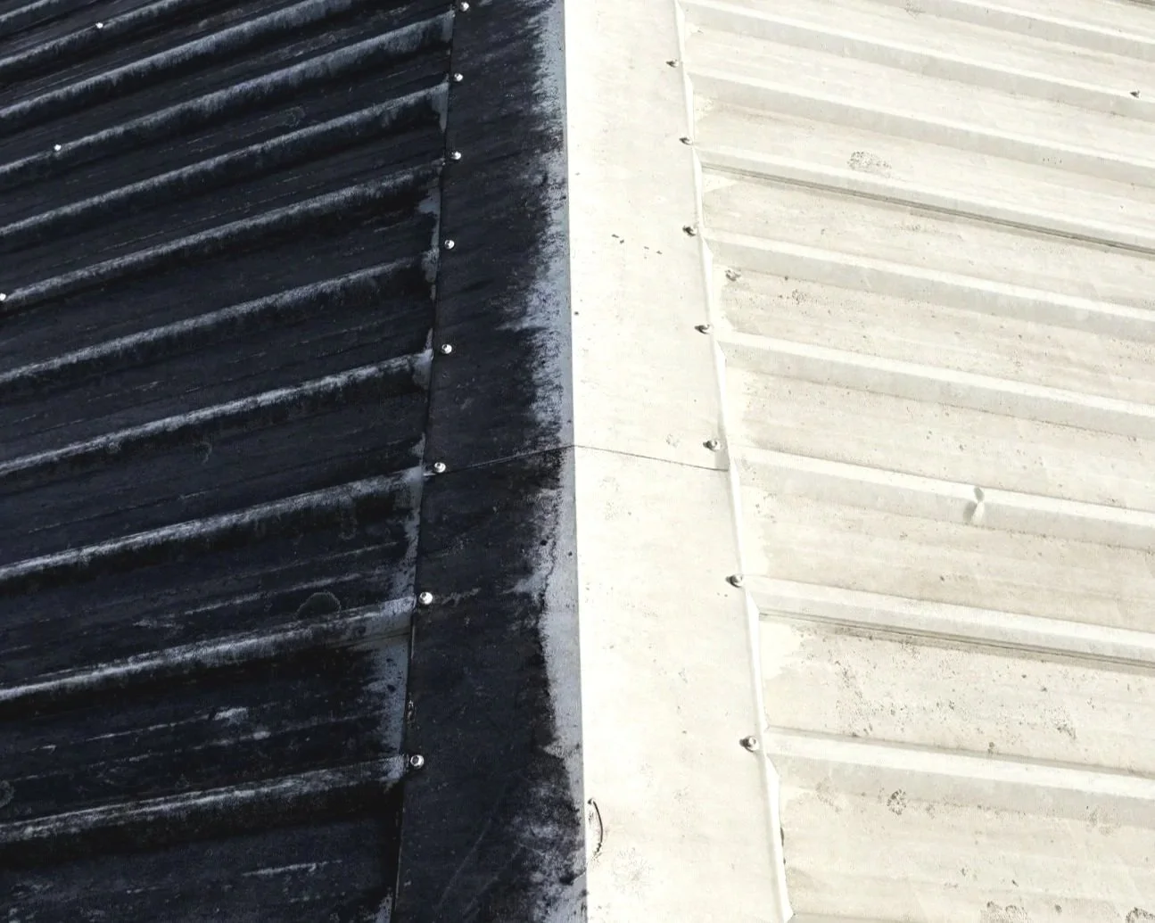 Close-up of two sections of a roof, one side with black, weathered corrugated metal and the other with white, cleaner corrugated metal, separated by a vertical line.