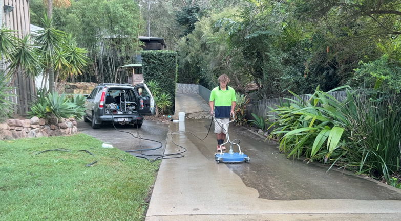 A man in a neon green shirt is cleaning a concrete sidewalk with a power washer, with a parked van nearby and lush greenery on both sides.