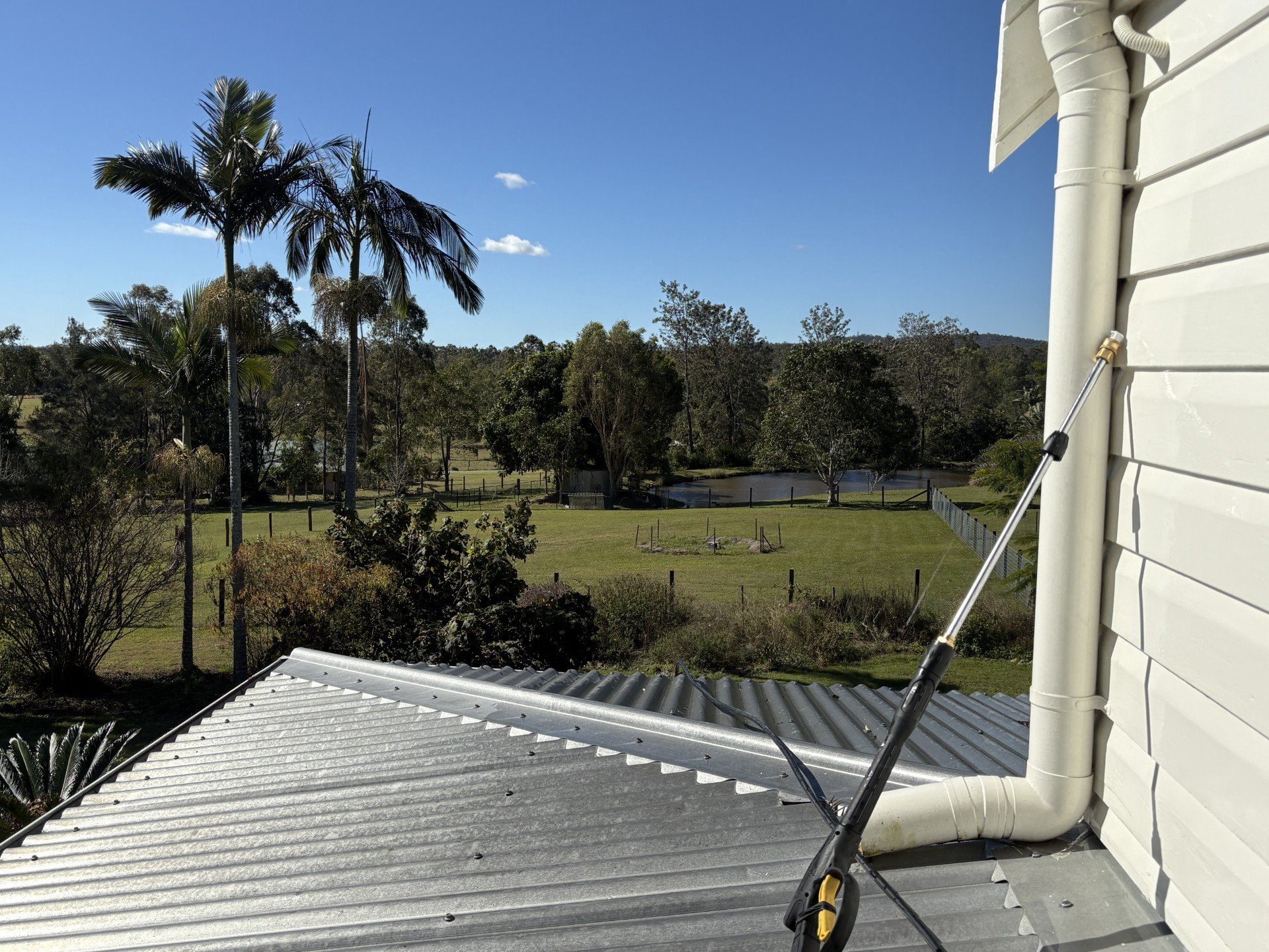 View from a rooftop showing a metal roof, white siding, a gutter and downspout, with a landscape of trees, grass, a pond, and a clear blue sky in the background.