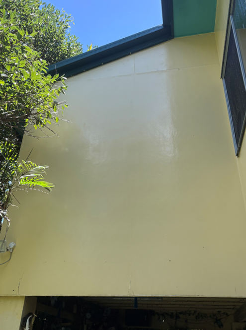 Exterior view of a yellow wall of a building with a window, sky, and trees.
