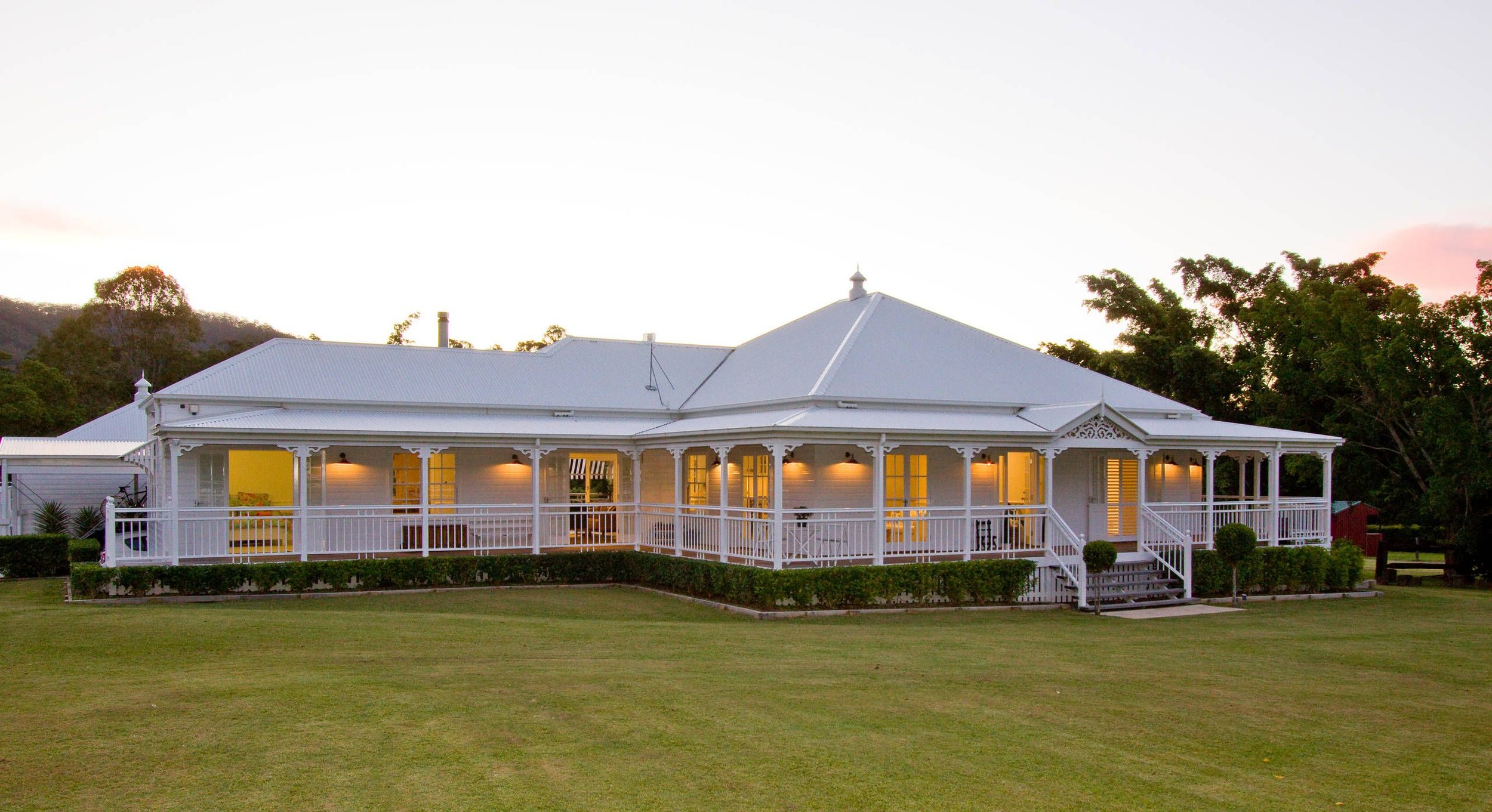 A large white house with a wraparound porch, glowing yellow lights inside, surrounded by a green lawn and trees at sunset.