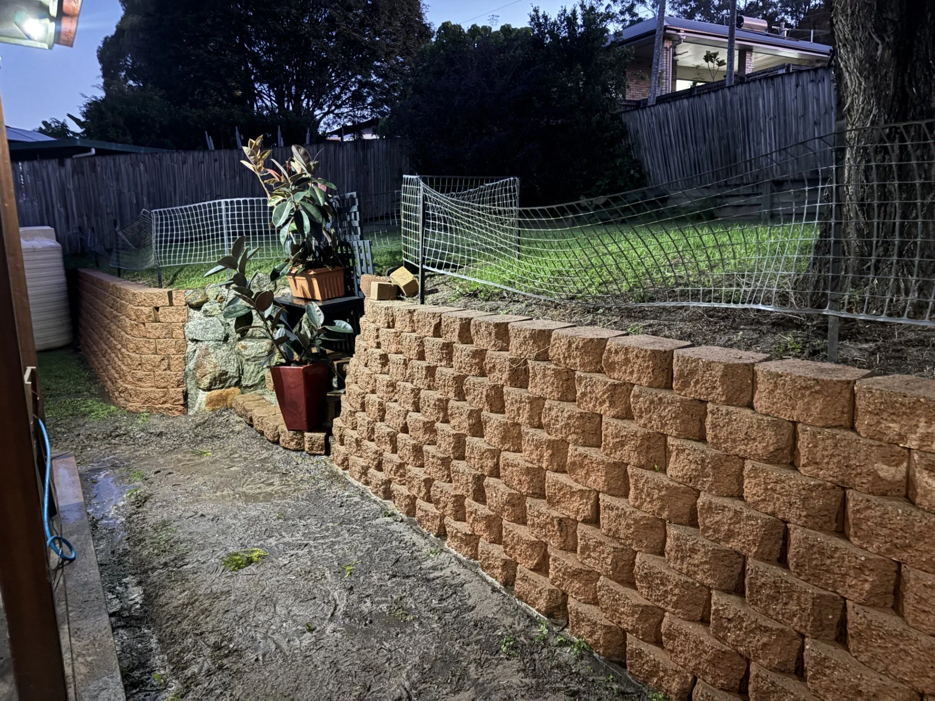 A backyard with a rock retaining wall made of reddish-brown bricks, with a dirt and concrete ground area and some potted plants on the wall. There are wire fences and a wooden fence on top of the retaining wall, with a grassy yard and trees beyond. A house with a balcony can be seen in the background.