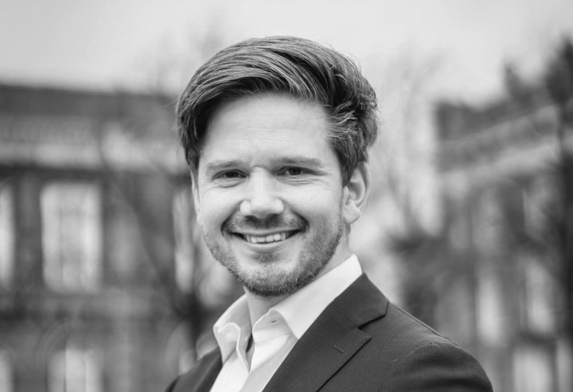 Foto van Gideon van Meijeren. Zwart-witportret van een glimlachende man met gestyled haar, gekleed in een pak en wit overhemd, buitenachtergrond. 


