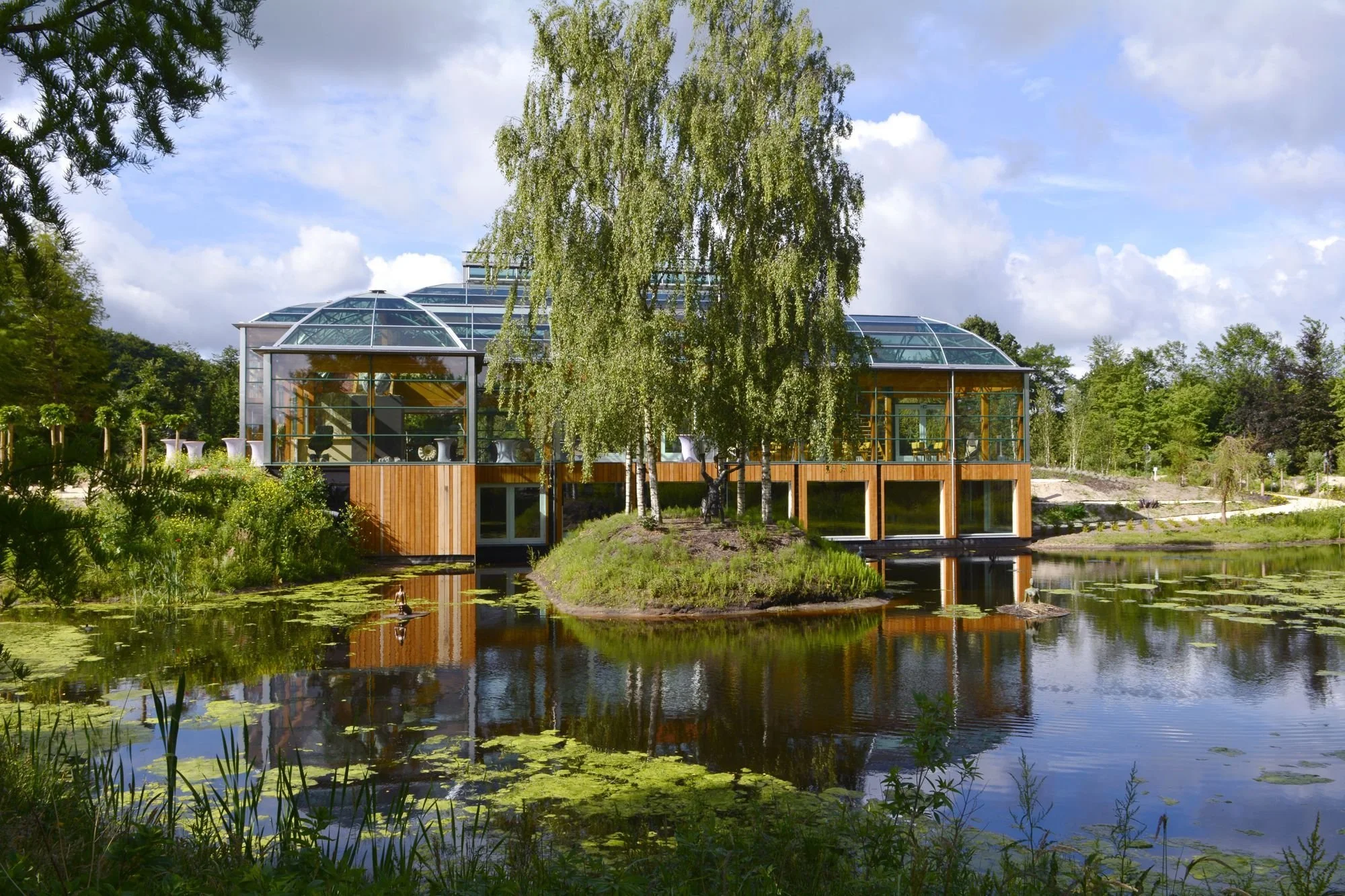 Een modern gebouw met grote glazen ramen en een koepelvormig glazen dak, omgeven door weelderig groen en een vijver met waterlelies, weerspiegeld in het water.