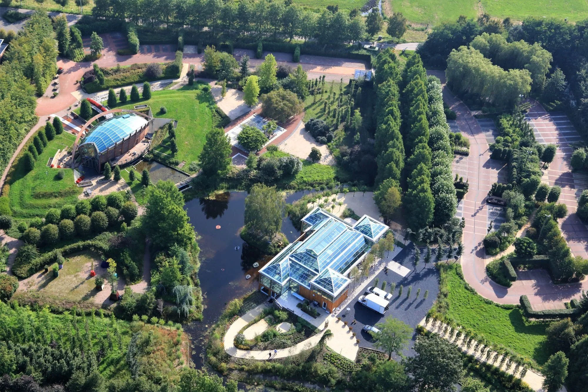 Luchtfoto van een weelderige groene botanische tuin met een glazen serre, vijver en aangelegde paden.