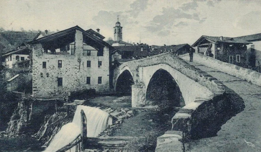 Fotografia in bianco e nero di un vecchio ponte di pietra su un fiume con alcune case circostanti, in stile rurale o storico.