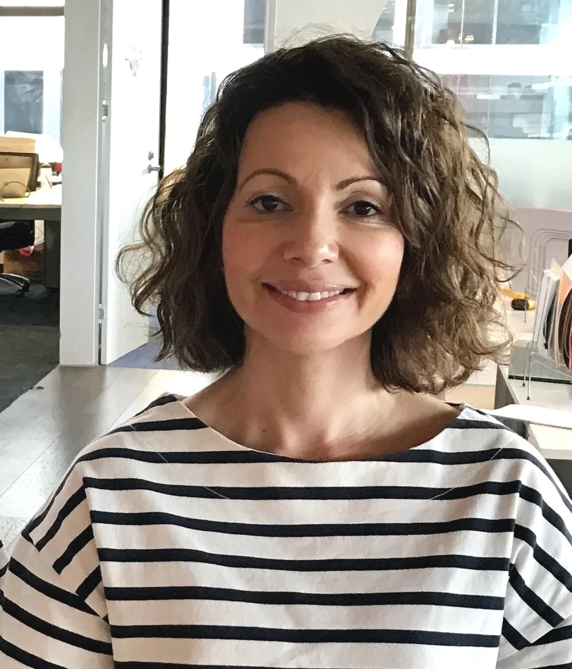 A woman with curly brown hair smiling, wearing a black and white striped shirt, in an office environment.