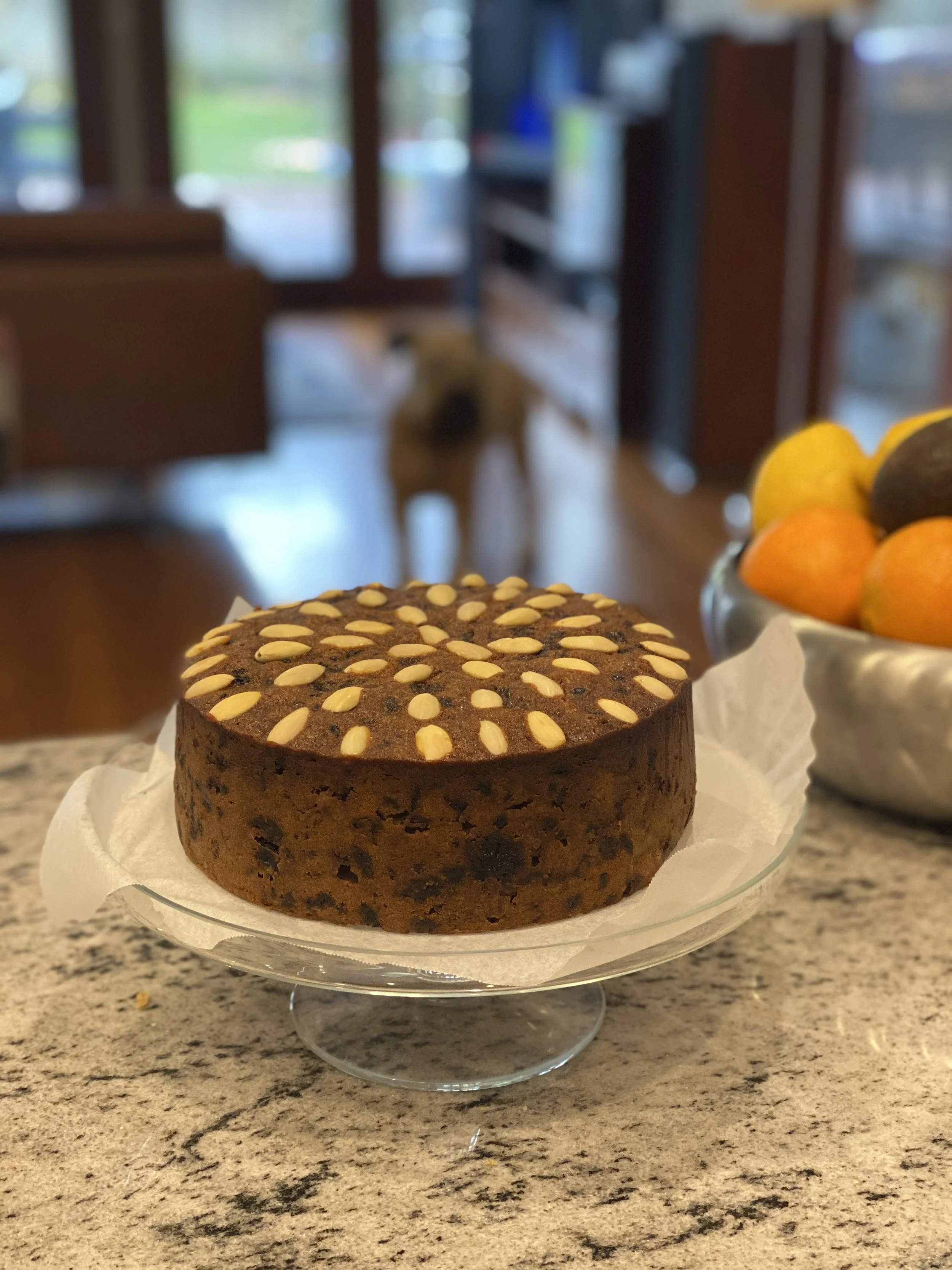 Chocolate cake topped with almond slices on a glass cake stand on a granite kitchen counter with a bowl of fruit in the background.
