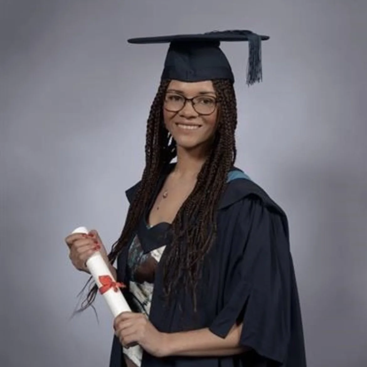 Woman with long braids, glasses, and a black graduation cap and gown holding a diploma with a red ribbon, smiling against a gray background.