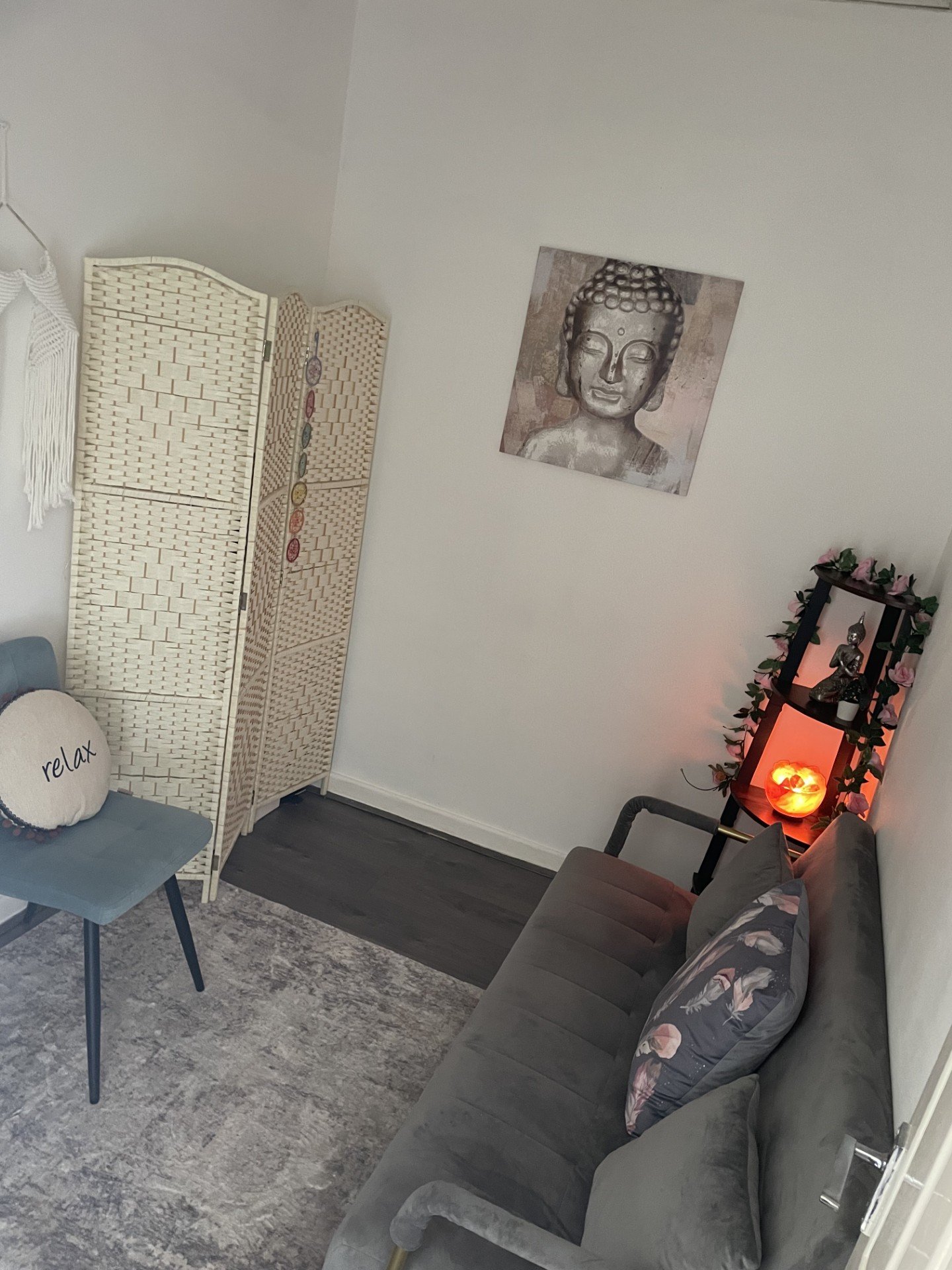 A cozy room with a gray sofa, decorative pillows, a black shelf with a Buddha statue, a candle and pink flowers, a wooden privacy screen, and wall art of a Buddha face.