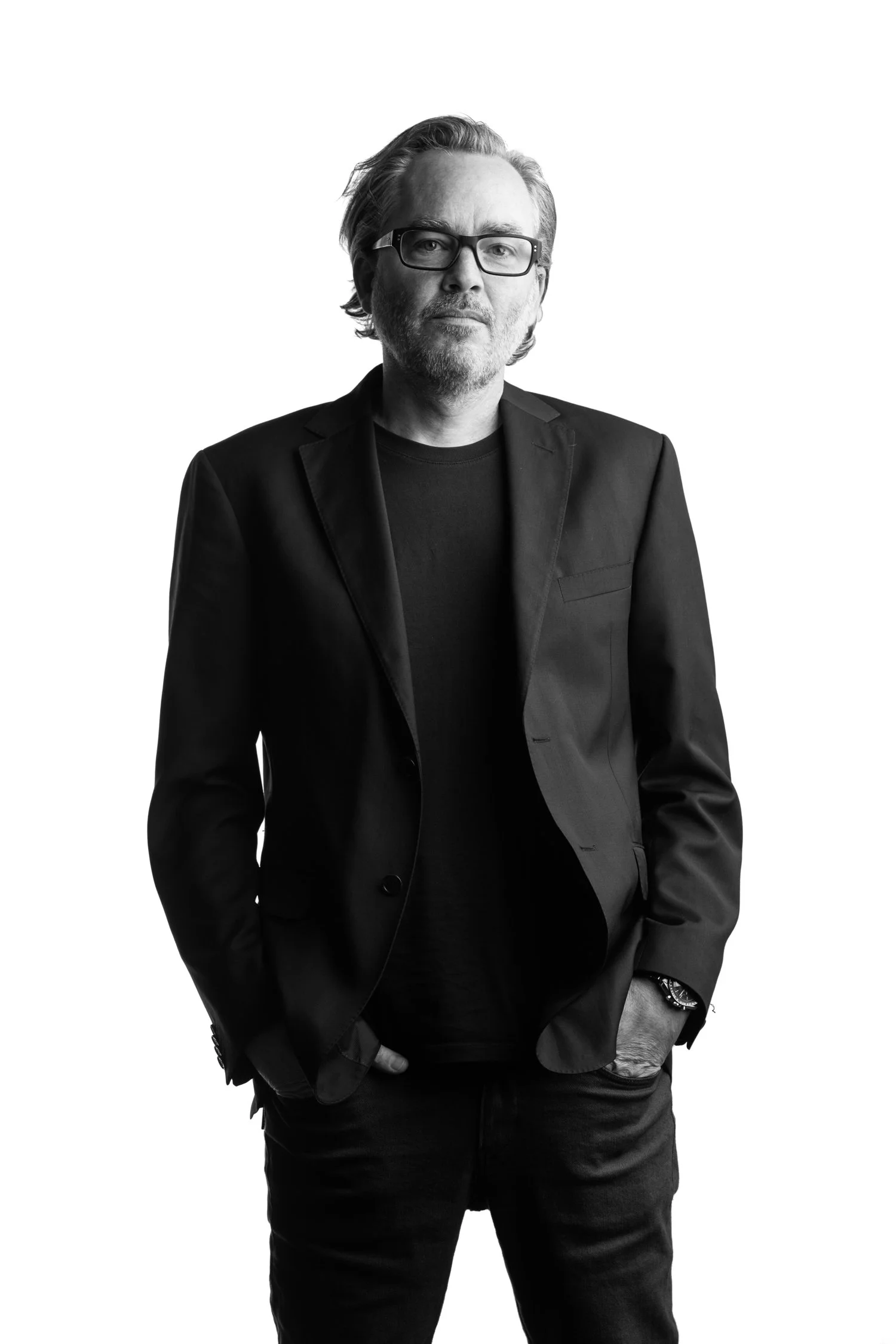 Black and white portrait of a man with glasses, wearing a black blazer and T-shirt, with hands in pockets, standing against a plain background.