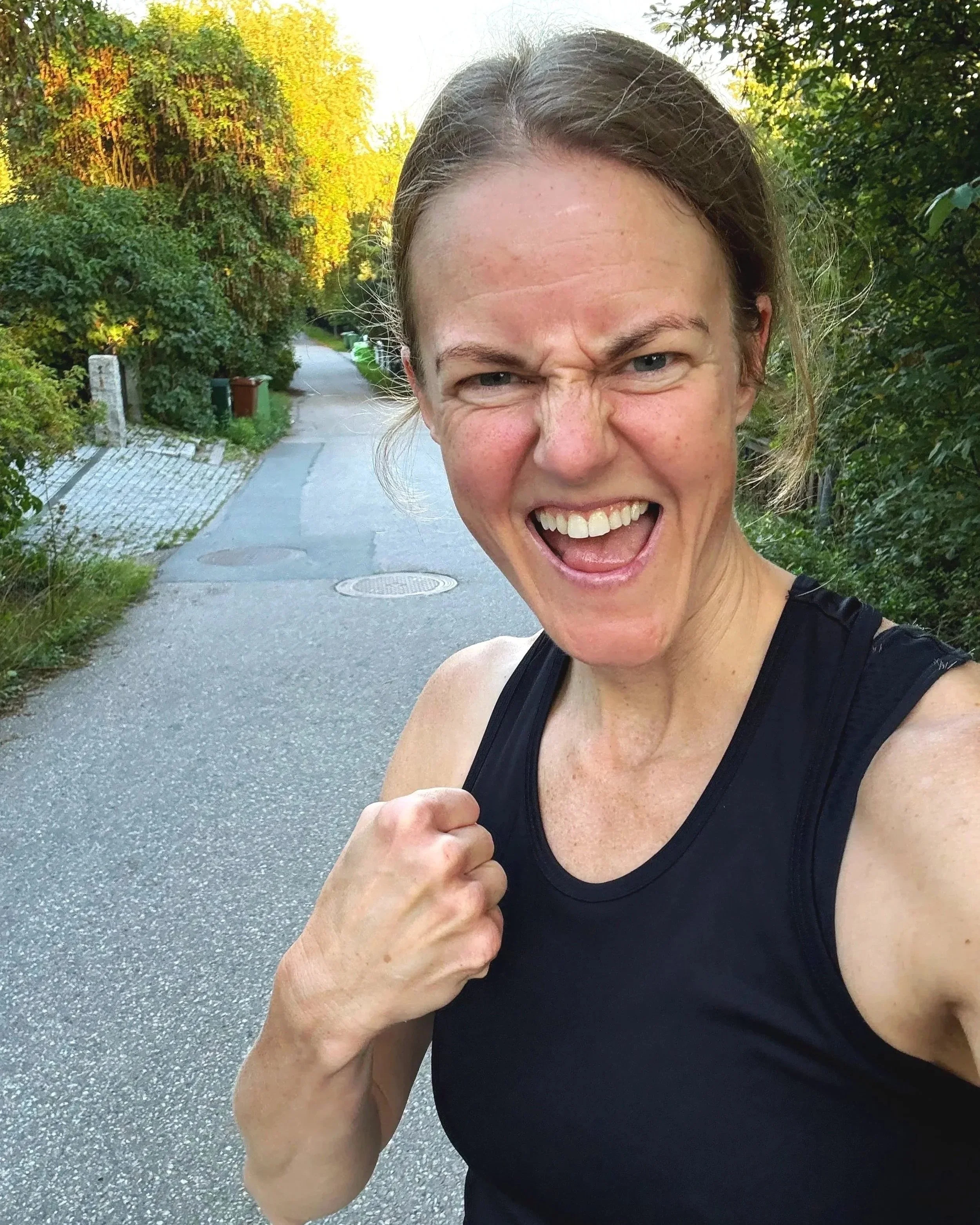 en kvinna som är ute och springer på en smal gata, skrattar och visar ett slagfält för näven, med gröna träd och trottoar i bakgrunden.