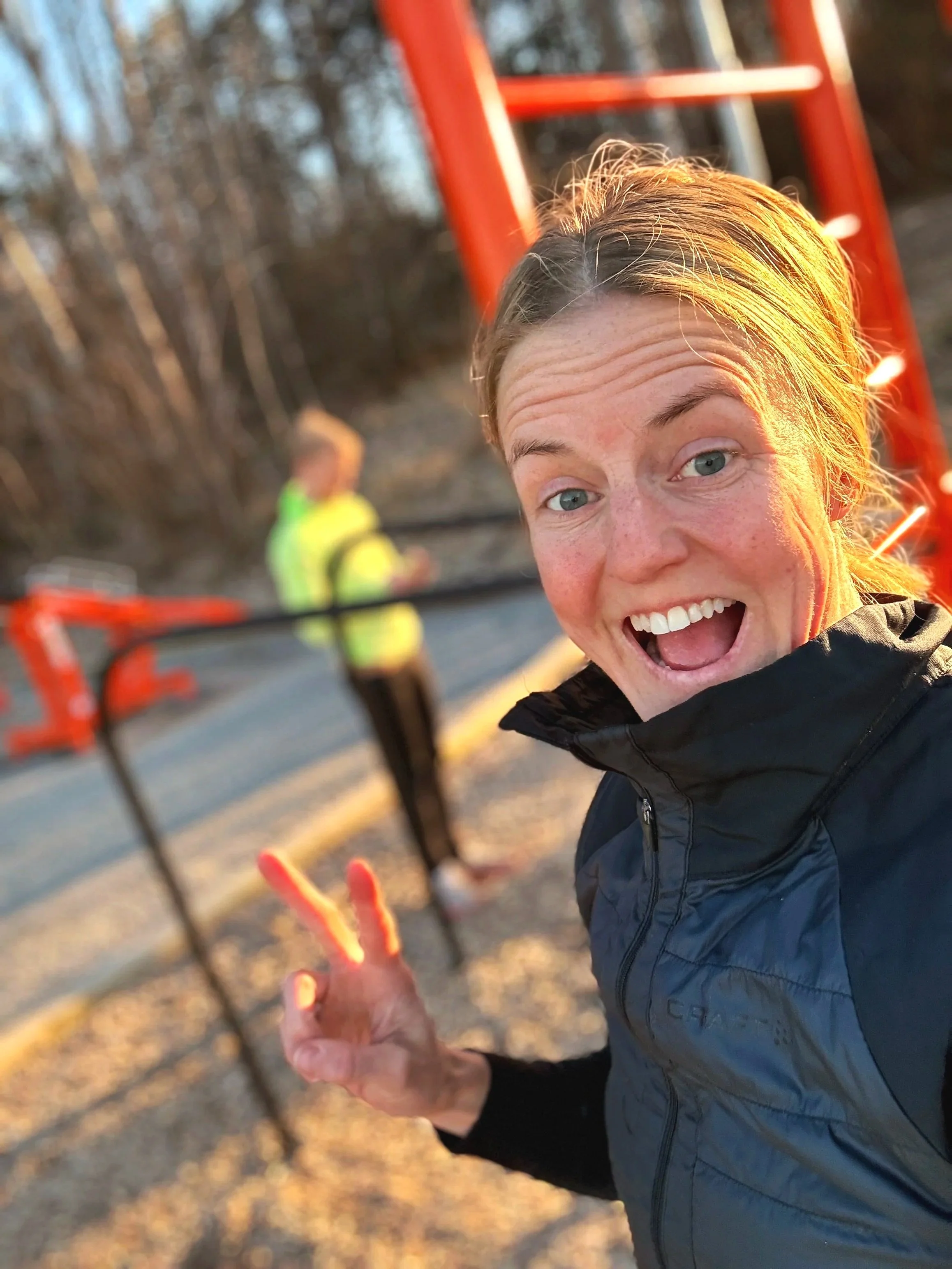 En glad kvinna framför en lekplats som gör en peace-tecken gest med handen. Bakom henne finns en person i en gul jacka och en orange klätterställning.