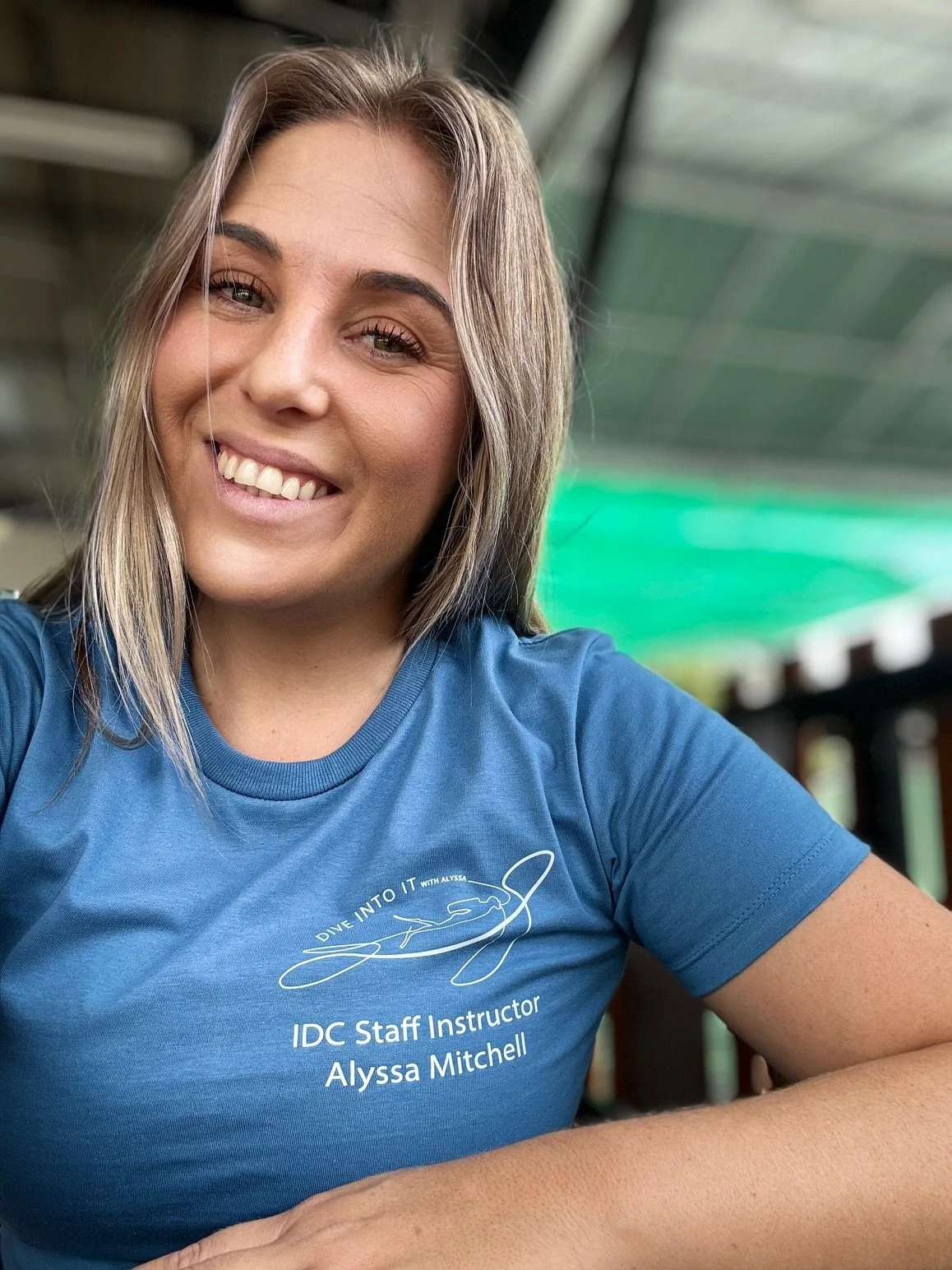 A woman smiling and taking a selfie, wearing a blue IDC Staff Instructor Alyssa Mitchell t-shirt, with a background of a green awning and outdoor setting.