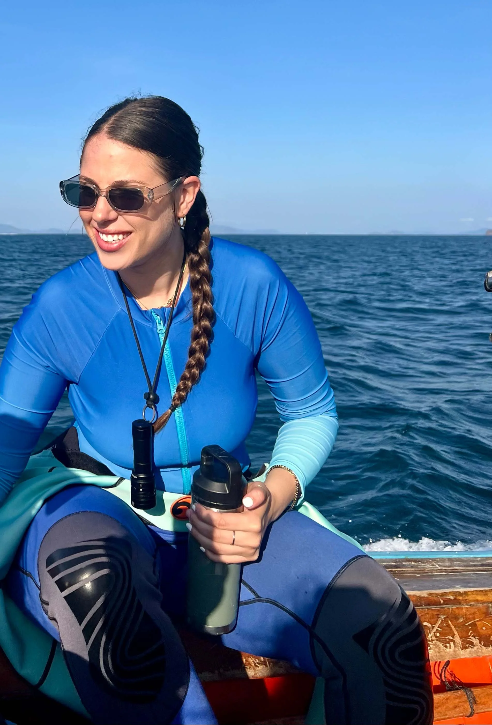 A woman with long braid, sunglasses, and a blue waterproof jacket sitting on a boat holding a water bottle, smiling, with open sea and clear sky in the background in a padi divemaster course in Thailand.