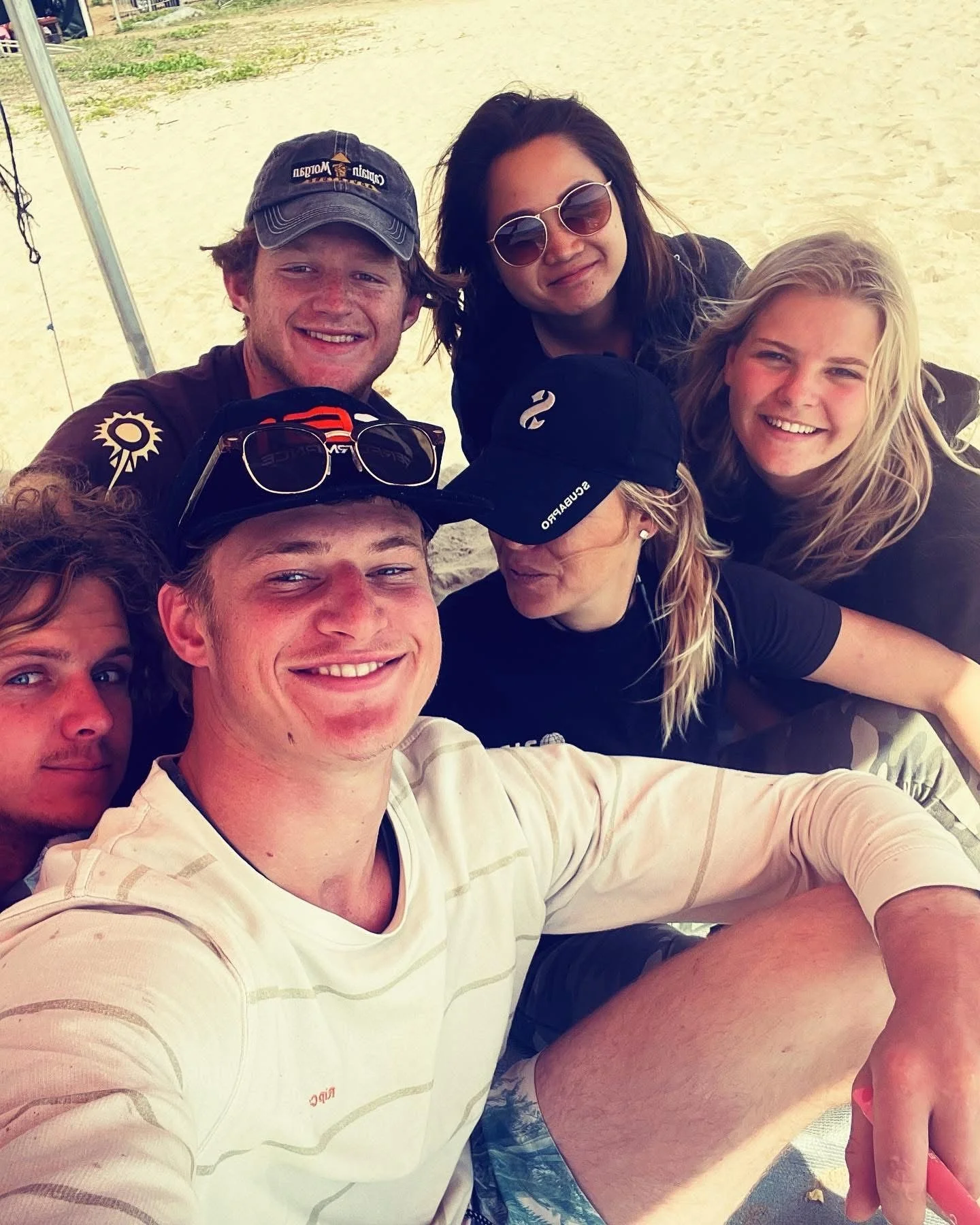 Group of seven young adults taking a selfie on the beach, smiling and wearing casual clothing and sunglasses, on a padi divemaster course.