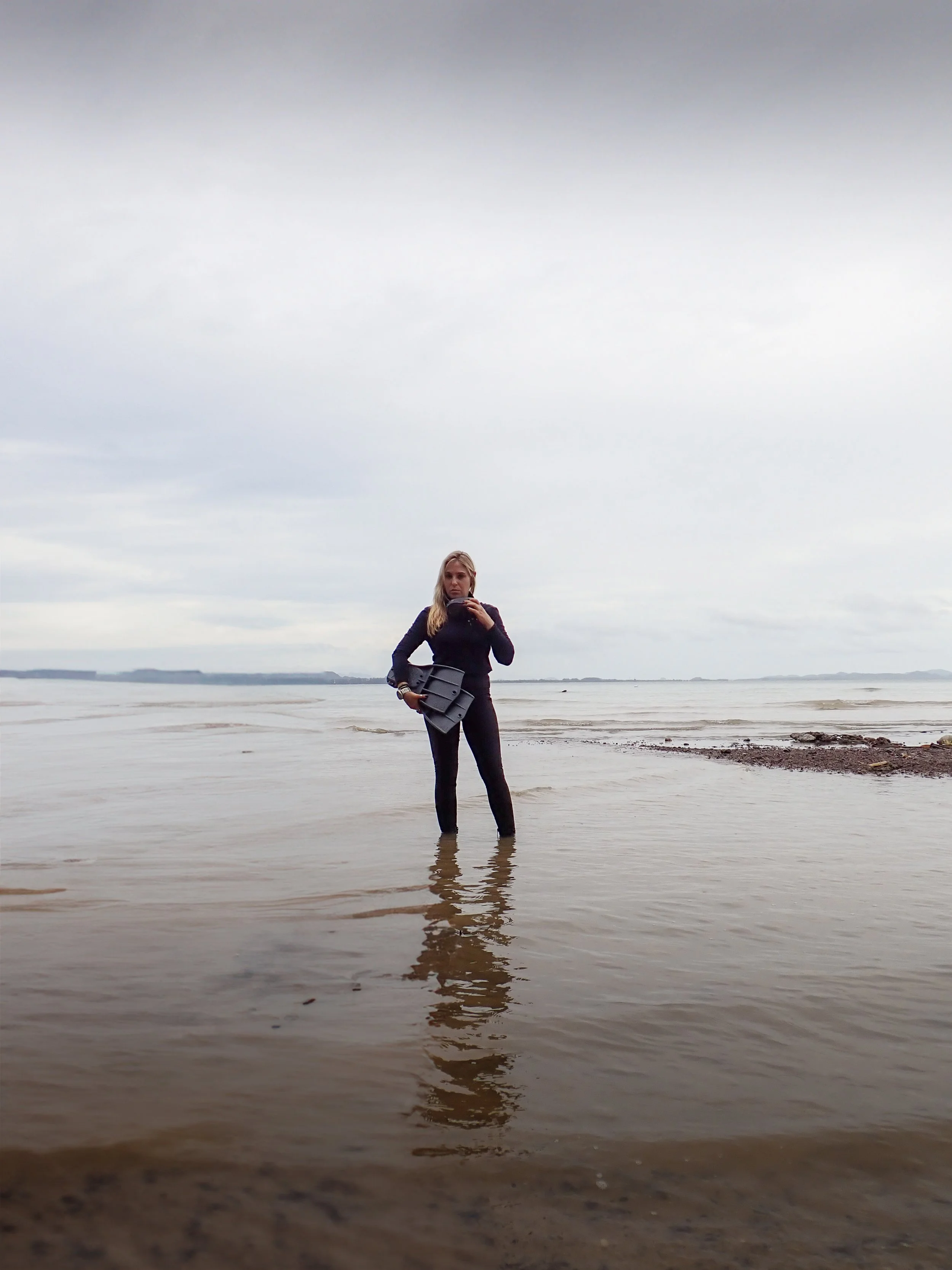 A woman in black standing in shallow water at the beach, holding a scuba gear, with an overcast sky in the background.