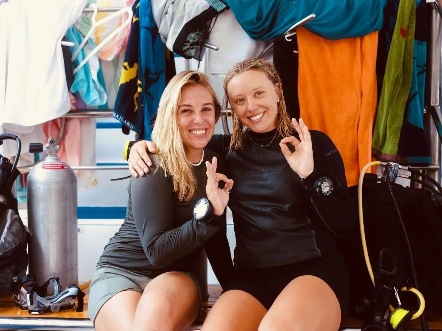 Two women in wetsuits posing happily with a sea diving mask and a tank, surrounded by hanging colorful towels and wetsuits in a diving shop or changing room.