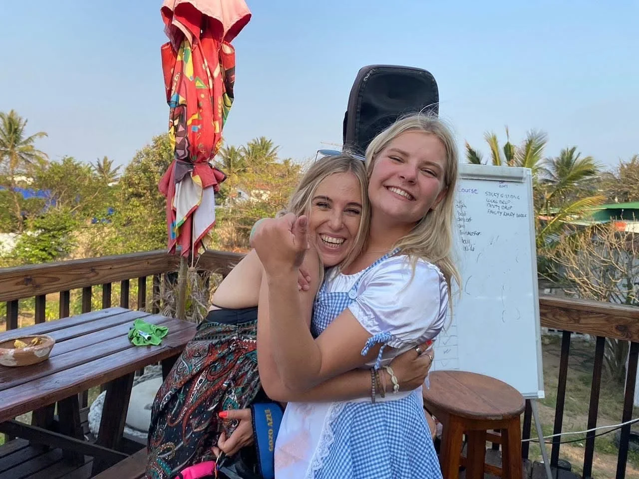 Two smiling women hugging on a wooden deck with umbrellas and a whiteboard in the background, trees, and a partly cloudy sky.
