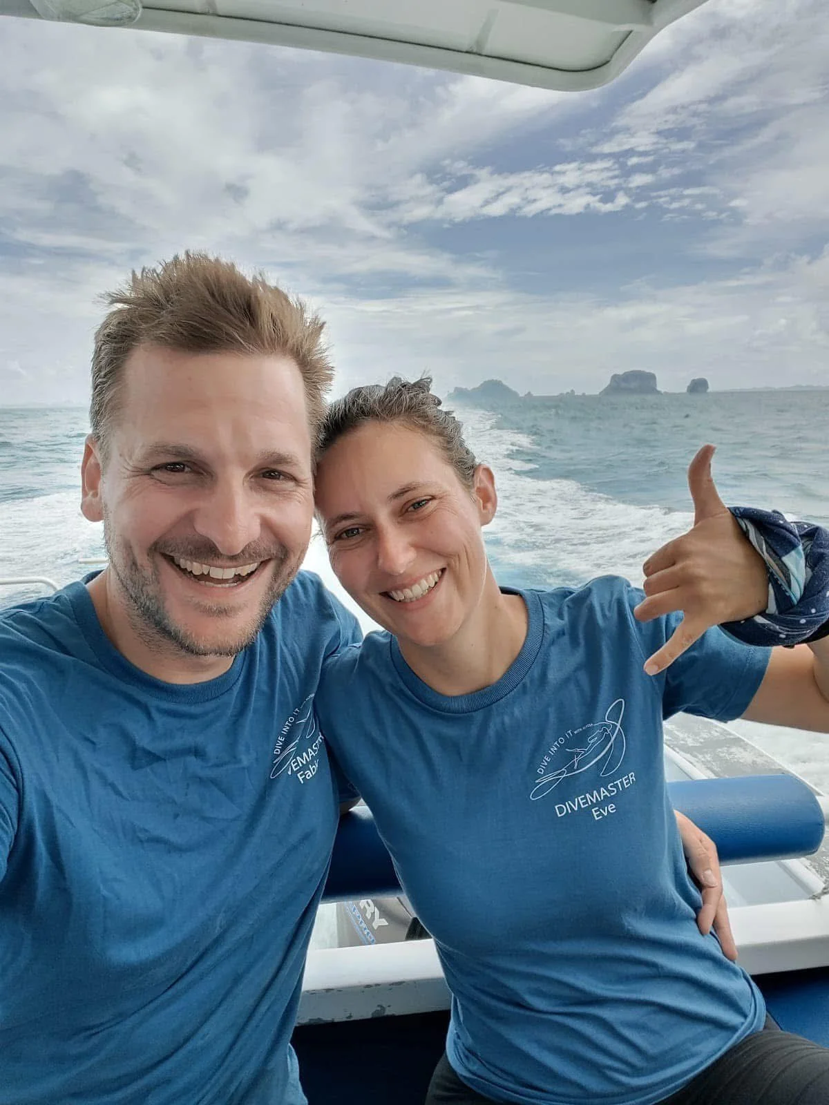 Two smiling people on a boat, taking a selfie with ocean and distant islands in the background. Both wearing blue t-shirts with diving-related logos in Thailand.
