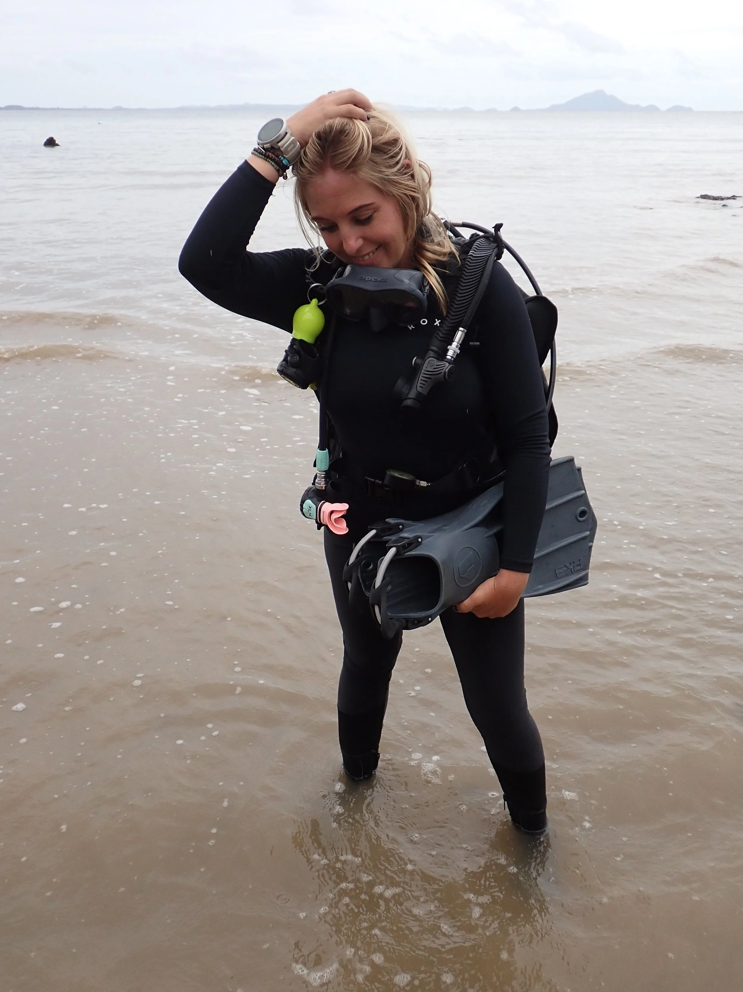 A woman standing in shallow ocean water, dressed in black wetsuit gear, smiling with hand on head, holding scuba diving fins, with a diving mask around her neck and diving equipment on her back.
