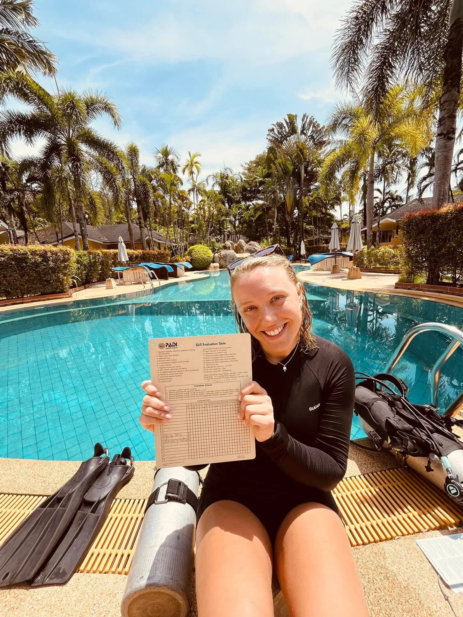 A smiling woman in a black long-sleeve swimsuit sitting by a swimming pool, holding a PADI skill evaluation slate, with scuba fins and gear beside her, surrounded by palm trees and sunny weather.